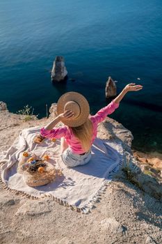 photo of a beautiful woman with long blond hair in a pink shirt and denim shorts and a hat having a picnic on a hill overlooking the sea.