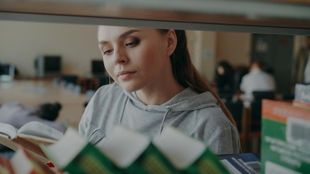 Young caucasian beautiful female student is standing near shelf with books in big lighty spacious library holding one, turning over pages and reading seriously