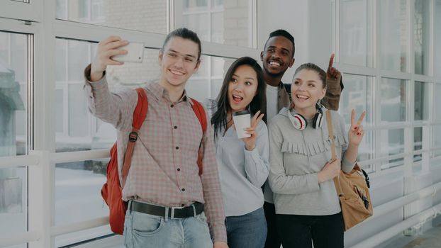 Group of four multi-ethnic positive male and female students are standing in wide white corridor of college. Hipster guy is holding smartphone making selfie of them all