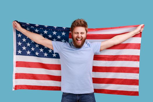 Portrait of handsome overjoyed bearded man holding USA flag and looking at camera with rejoicing look, celebrating national holiday. Indoor studio shot isolated on blue background.