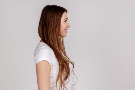Side view portrait of beautiful woman with charming smile, standing and looking at ahead, expressing positive emotions, wearing white T-shirt. Indoor studio shot isolated on gray background.