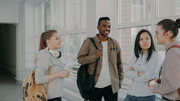 Young beautiful caucasian female student is standing in front of her three multi-ethnic groupmates discussing something together in positive way near window in wide hallway.