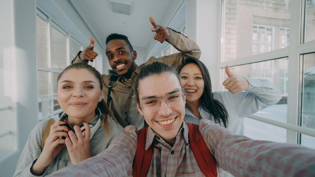 Point of view of four positive multi-ethnic group of friends talking selfie photos holding smartphone and having fun laughing while standing in corridor of university.