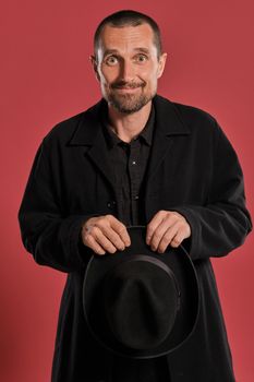 Close-up studio shot of an adult fellow with beard and mustache, wears black jacket, holding a hat in his arms and looking at the camera while posing against a red background with copy space. Sincere emotions concept.