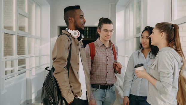 Multi-ethnic group of four students standing in wide lighty glassy hall in college talking to each other in positive way, Guys and girls are smiling ant laughing dinking coffee