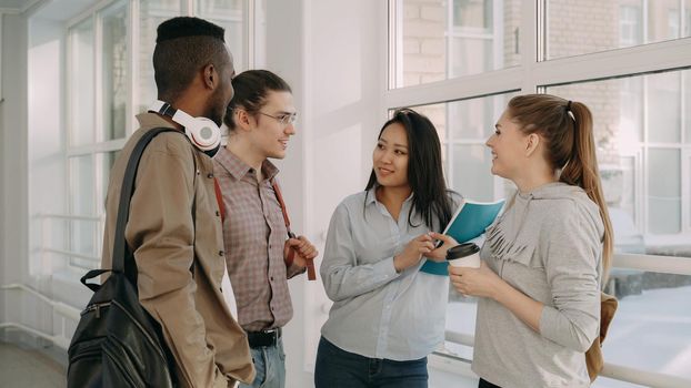 Four multi-ethnic studens are standing in big white glassy spacious hall in college talking to each other about study in positive way. They are smiling positively and laughing