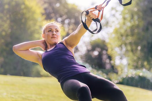 attractive young woman training fitness with suspension straps, during functional workout in a sunny outdoor park in the morning