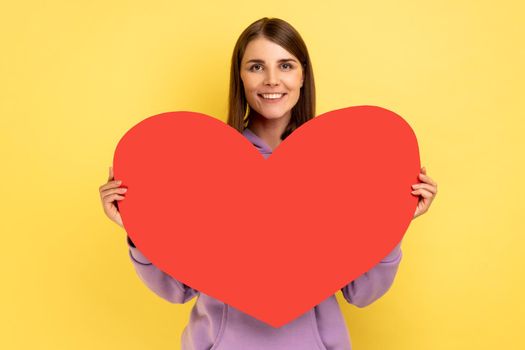 Portrait of happy cheerful woman holding in hands big red paper heart and smiling at camera, love and romance, wearing purple hoodie. Indoor studio shot isolated on yellow background.