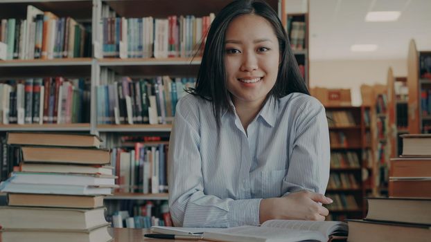 Portrait of young beautiful asian female student sitting at table with huge piles of textbooks and copybook in front of her in library looking at camera. She is smiling positively.