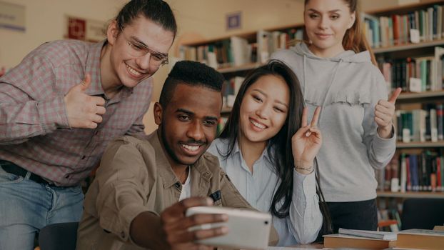 Group of international students have fun smiling and making selfie photos on smartphone camera at university library indoors. Cheerful friends have rest while preapre project together