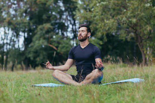 A young man with a beard sitting in the park on a mat meditates, performs exercises to improve breathing, active lifestyle