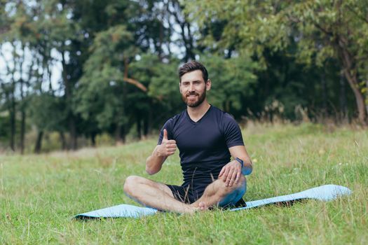 A young man with a beard sitting in the park on a mat meditates, performs exercises to improve breathing, active lifestyle