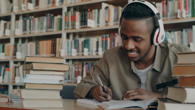 Handsome african american male student wearing big white headphones is listening to music sitting at table in big spacious library writing lecture surrounded by books