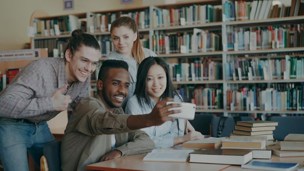 Group of international students have fun smiling and making selfie photos on smartphone camera at university library indoors. Cheerful friends have rest while prepare for examination