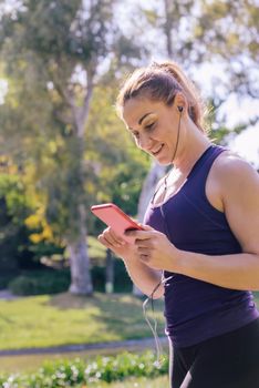 vertical photo of an attractive blonde sports woman listening music before workout outdoors at park, fitness accessories