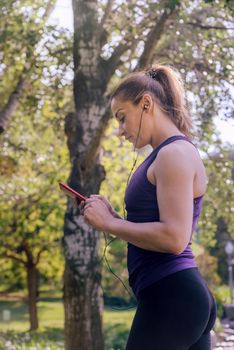 vertical photo of an attractive blonde sporty woman checking her phone app before running outdoors at park, fitness accessories