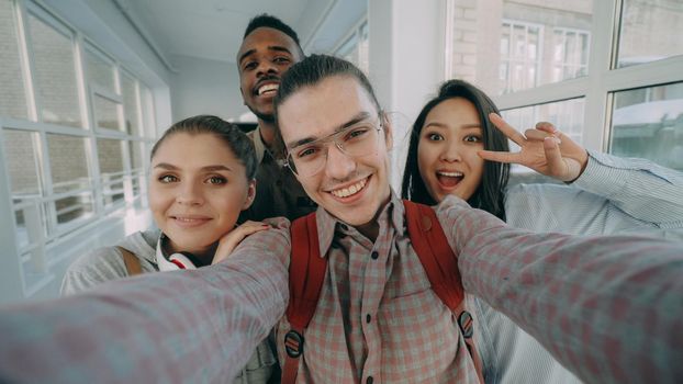 Point of view of four positive cheerful attractive multi-ethnic friends talking selfie photos holding smartphone and having fun laughing while standing in corridor of university.