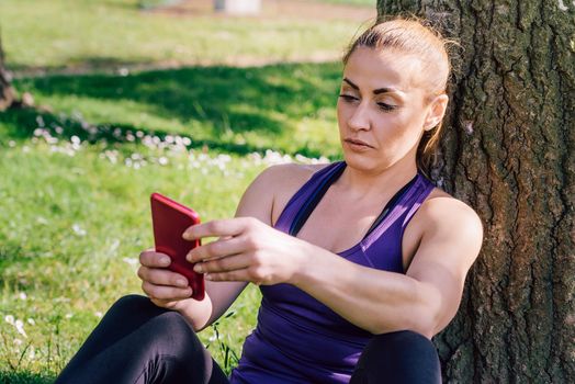 fitness woman taking a workout rest sitting next to a tree for texting on her phone, healthy modern lifestyle and sport concept