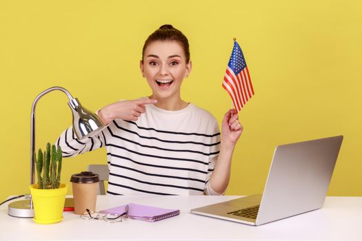 Portrait of young excited delighted woman office manager holding flag of USA and looking at camera with amazement and happiness. Indoor studio studio shot isolated on yellow background.