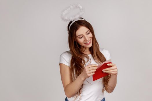 Portrait of charming pleased woman with nimbus over head holding red envelope, reading romantic letter with smile, wearing white T-shirt. Indoor studio shot isolated on gray background.