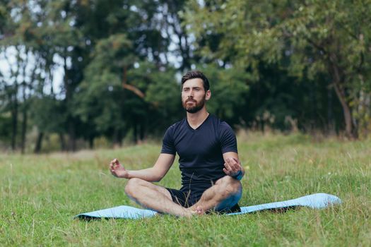 A young man with a beard sitting in the park on a mat meditates, performs exercises to improve breathing, active lifestyle