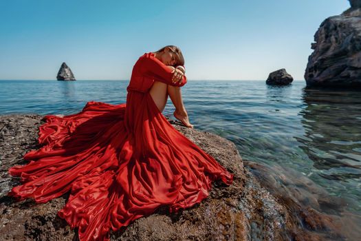 Beautiful sensual woman in a flying red dress and long hair, sitting on a rock above the beautiful sea in a large bay