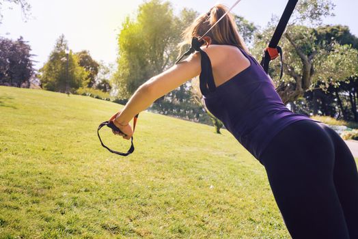 young beautiful blonde woman using suspension straps, exercise routine outdoors at the evening in a sunny park