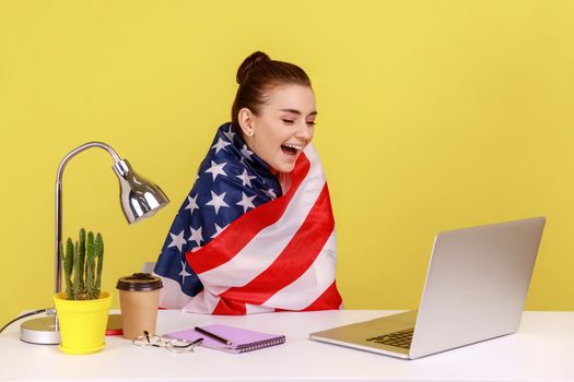 Happy woman employee sitting wrapped in USA flag, shouting for joy in office workplace, celebrating labor day or US Independence day. Indoor studio studio shot isolated on yellow background.