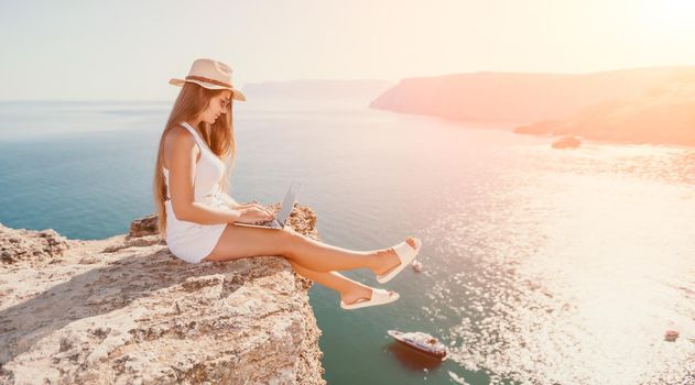 Successful business woman in yellow hat working on laptop by the sea. Pretty lady typing on computer at summer day outdoors. Freelance, travel and holidays concept.