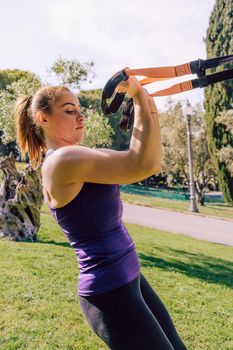 vertical photo of a strong young woman training biceps with suspension fitness straps, she is outdoors during functional workout