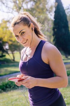 vertical photo of an attractive blonde athlete woman listening music before her training outdoors at park, fitness accessories