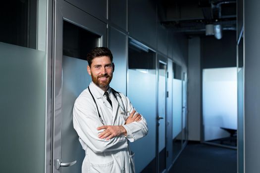 Friendly doctor smiling with crossed arms, man in medical gown, near ward with patient, looking at camera having fun