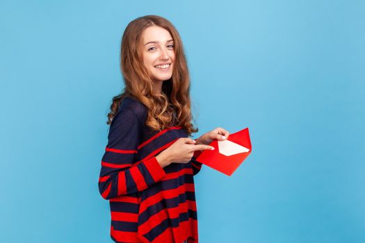I got love letter on Valentine's day. Woman in striped casual style sweater, pointing letter in red envelope, holding greeting card and smiling joyfully.Indoor studio shot isolated on blue background.