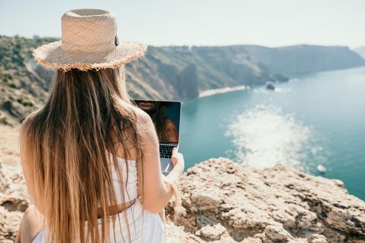 Successful business woman in yellow hat working on laptop by the sea. Pretty lady typing on computer at summer day outdoors. Freelance, travel and holidays concept.