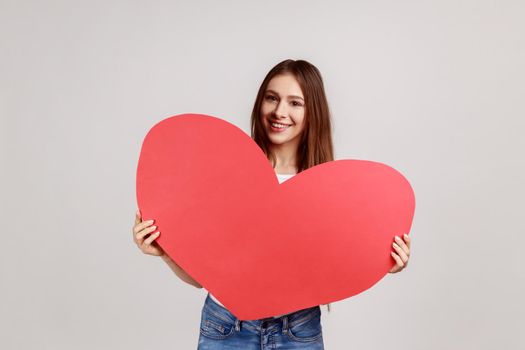 Happy Valentine's day, Portrait of beautiful woman holding red heart, big greeting card, mock up for romantic congratulation, wearing white T-shirt. Indoor studio shot isolated on gray background.