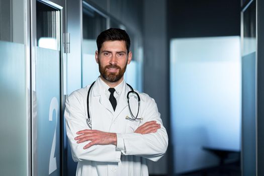 Friendly doctor smiling with crossed arms, man in medical gown, near ward with patient, looking at camera having fun