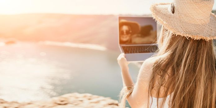 Successful business woman in yellow hat working on laptop by the sea. Pretty lady typing on computer at summer day outdoors. Freelance, travel and holidays concept. Back view.