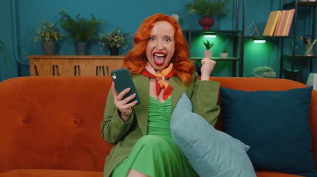 Excited happy redhead woman use mobile smartphone typing browsing say Wow yes found out great big win good news celebrate doing winner gesture. Young ginger girl at home in living room sitting on sofa