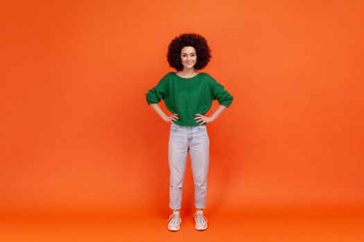 Full length portrait of positive woman with Afro hairstyle wearing green casual style sweater standing with hands on hips and toothy smile. Indoor studio shot isolated on orange background.