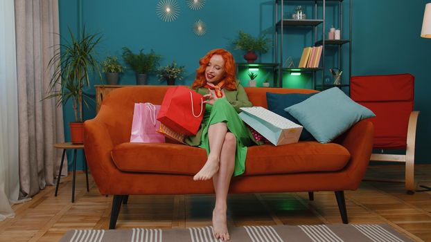 Happy redhead young woman shopaholic consumer in green dress came back home after shopping sale with bags at modern apartment. Satisfied ginger girl received parcels from online order in living room
