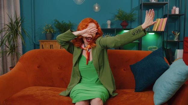 Trendy cheerful positive redhead young woman having fun dancing and moving to rhythm, dabbing raising hands, making dub dance gesture at modern home apartment. Ginger girl in living room on sofa