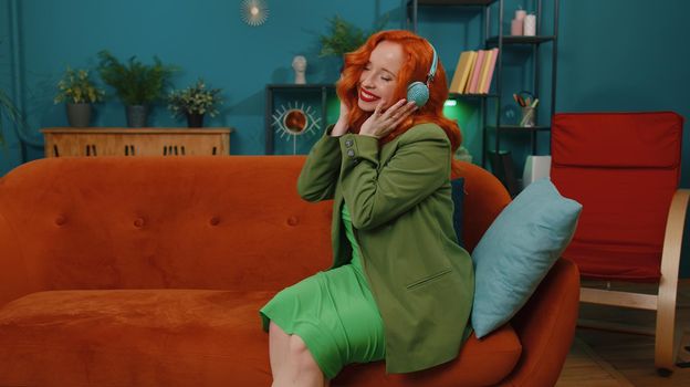 Happy relaxed overjoyed redhead girl in wireless headphones dancing on couch at home choosing listening favorite energetic disco rock n roll music in smartphone entertaining. People weekend activities