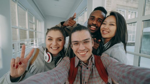 Point of view of four positive multi-ethnic group of friends talking selfie photos holding smartphone and having fun laughing while standing in corridor of university.