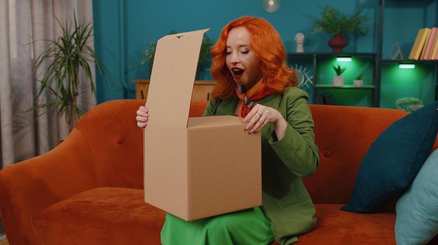 Happy redhead woman unpacking delivery parcel sitting at home. Smiling satisfied female ginger girl shopper, online shop customer opening cardboard box receiving purchase gift by fast postal shipping