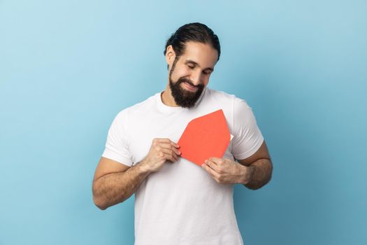 Portrait of smiling man wearing white T-shirt standing, holding embracing red envelope and keeping eyes closed, enjoying, being touched. Indoor studio shot isolated on blue background.