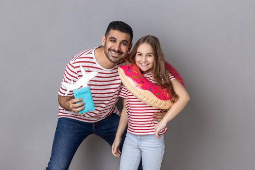 Portrait of happy dad and his daughter holding rubber ring and passport with paper plane, rejoicing their vacation abroad to the sea. Indoor studio shot isolated on gray background.
