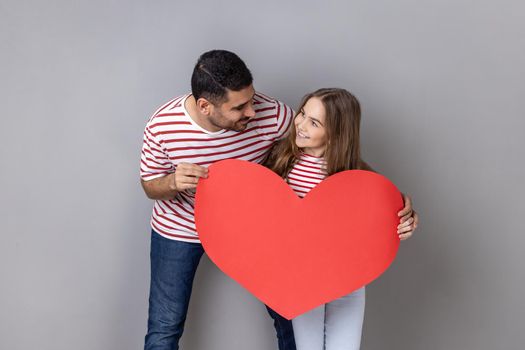 Portrait of loving father and charming cute daughter in striped T-shirts holding big red hear, looking at each other with love and gentle. Indoor studio shot isolated on gray background.