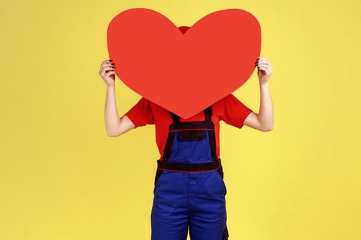 Portrait of optimistic worker woman hiding her face behind big red heart, expressing romantic feelings, wearing overalls and red cap. Indoor studio shot isolated on yellow background.