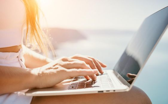 Successful business woman in yellow hat working on laptop by the sea. Pretty lady typing on computer at summer day outdoors. Freelance, travel and holidays concept.