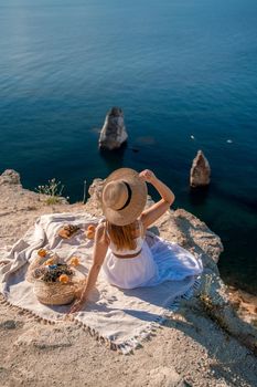 Street photo of a beautiful woman with dark hair in a white dress and hat having a picnic on a hill overlooking the sea.